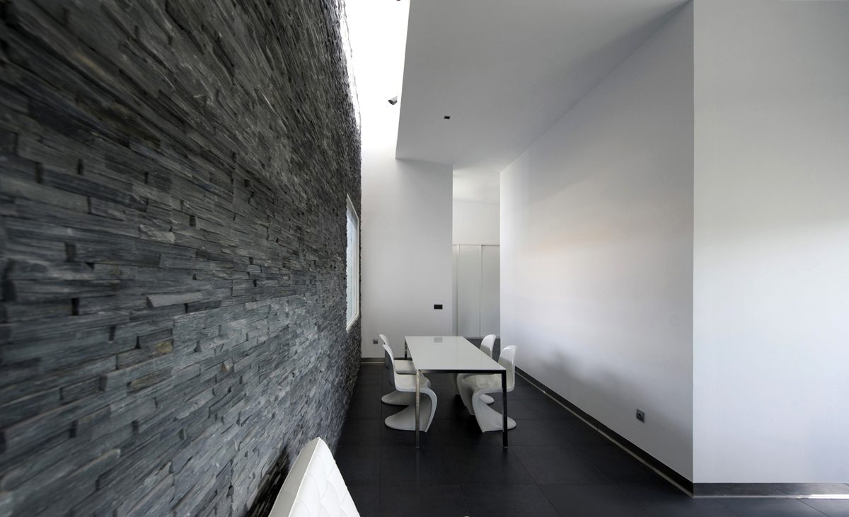 AQSO arquitectos office. El interior de la sala de estar tiene un muro de piedra natural de color negro iluminado por un lucernario. La mesa de comedor está rodeada de las sillas apilables diseñadas por Vener Panton.